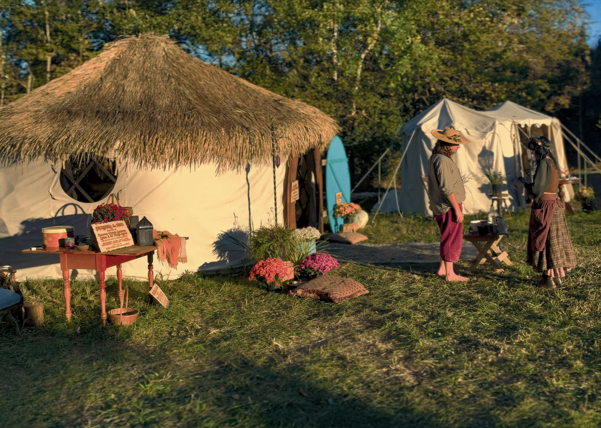 A finished ThatchTop Burrow in daylight — thatched roof and round door — set up among the festival tents with flowers out front