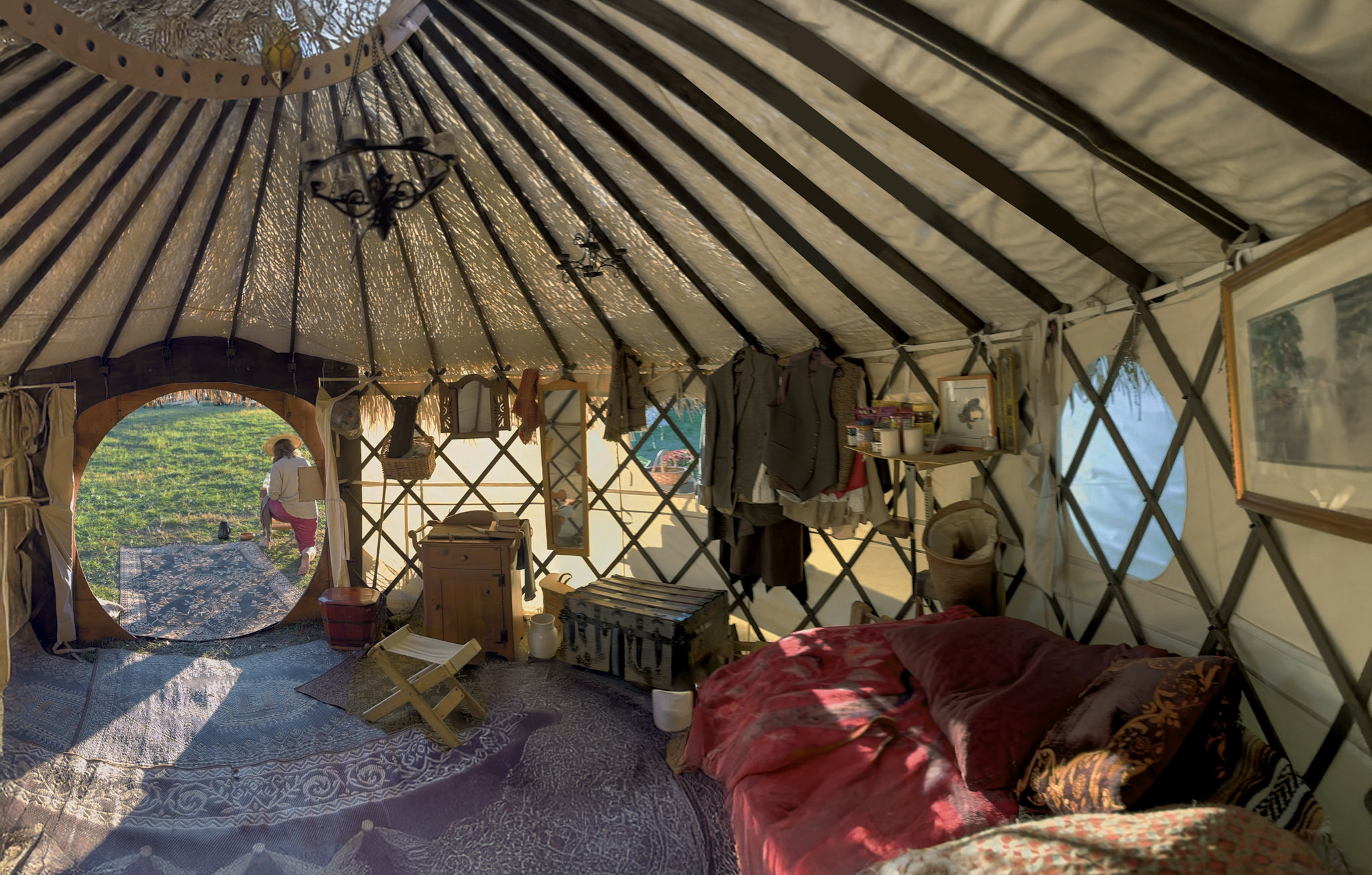 The daylit interior of a ThatchTop Burrow — lattice walls hung with belongings, a made bed, and the round door open onto a green field