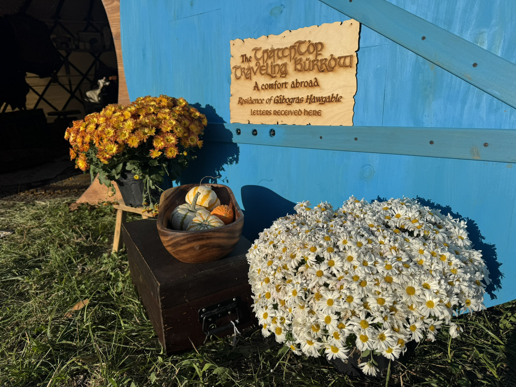 A hand-carved wooden sign reading 'The ThatchTop Traveling Burrow' beside the round door, framed by autumn flowers and gourds